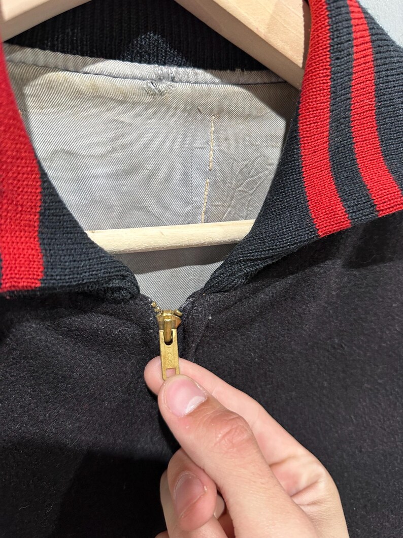 Op de afbeelding: Close-up van een zwarte jas met een rood-zwarte gestreepte kraag. De jas heeft een gouden rits en een grijze voering. De jas hangt aan een houten kleerhanger.