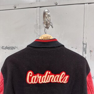 Op de afbeelding: Zwarte varsity jas met rode leren mouwen. Het woord "Cardinals" is in rood script op de rug geborduurd. De kraag is zwart met rood-witte strepen.