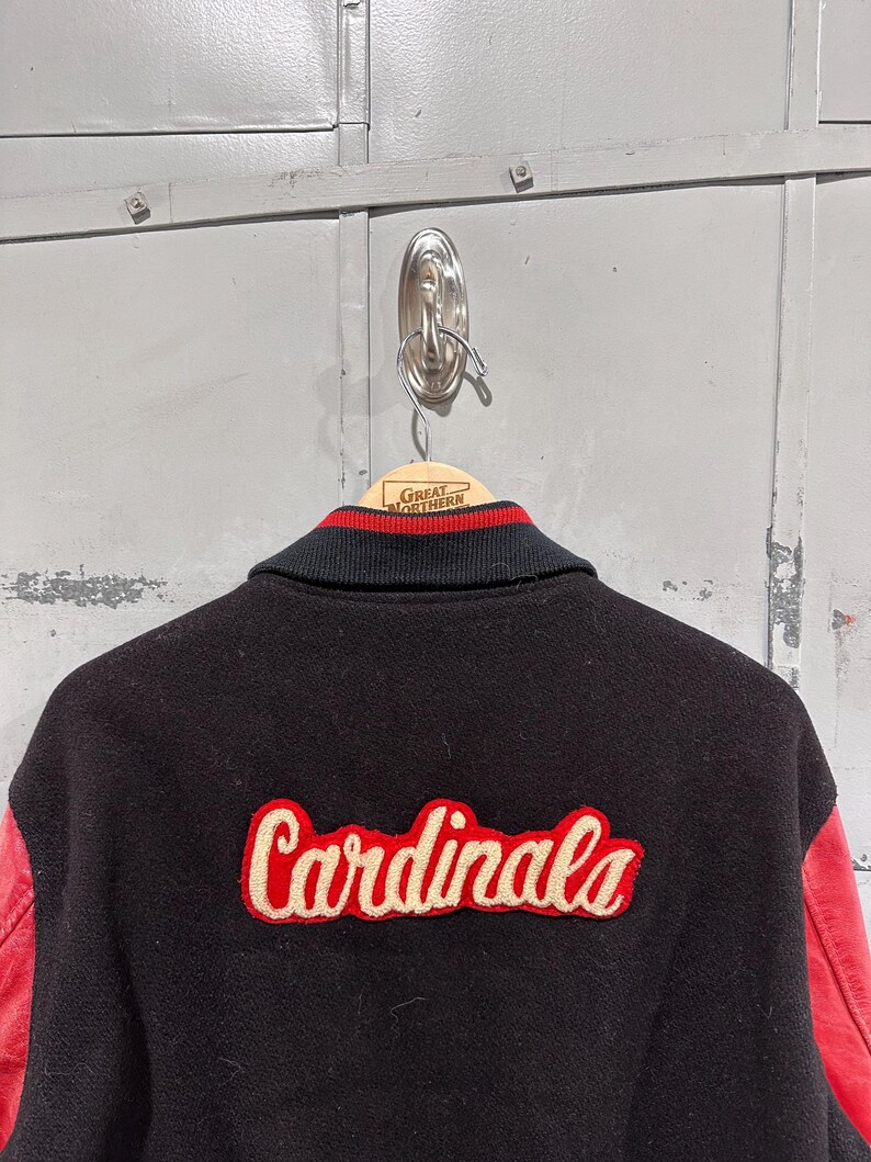 Op de afbeelding: Zwarte varsity jas met rode leren mouwen. Het woord "Cardinals" is in rood script op de rug geborduurd. De kraag is zwart met rood-witte strepen.