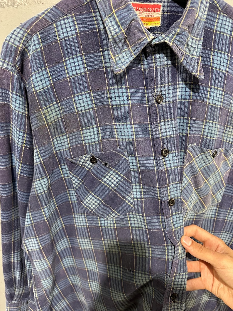 Pu&ograve; includere: Una camicia a maniche lunghe con bottoni caratterizzata da un motivo a quadri blu e blu navy con sottili linee gialle. La camicia presenta un colletto a punta, due tasche sul petto con bottoni e un'etichetta sul collo con la scritta FLANNELCRAFT 100% COTTON.