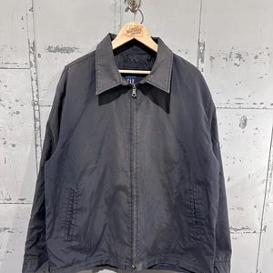 May include: A dark gray Gap jacket with a full zipper closure and a collared neckline. The jacket has two front pockets and is hanging on a wooden hanger. The hanger has the text "Great Northern Vintage".