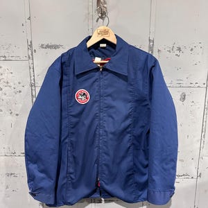 Puede incluir: Chaqueta azul marino con cremallera y cuello, colgada de una percha de madera. La chaqueta presenta un parche circular con texto y un gráfico. La cremallera tiene un acento rojo. La chaqueta se muestra sobre un fondo gris texturizado.