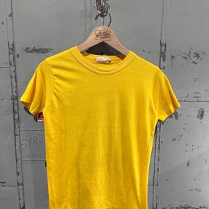 May include: A bright yellow, short-sleeved t-shirt hangs on a wooden hanger. The shirt has a crew neck and a simple, classic design. The hanger has a tag that reads "Great Northern Vintage". The shirt is displayed against a gray metal background.