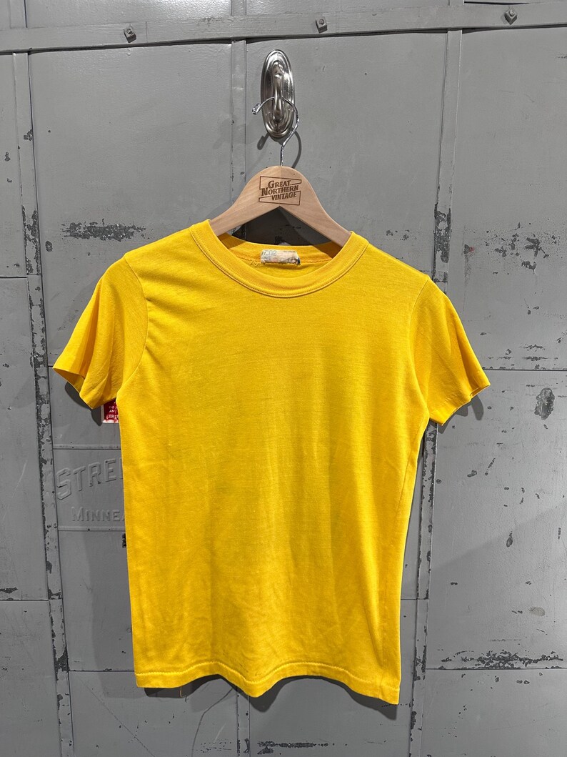 May include: A bright yellow, short-sleeved t-shirt hangs on a wooden hanger. The shirt has a crew neck and a simple, classic design. The hanger has a tag that reads "Great Northern Vintage". The shirt is displayed against a gray metal background.