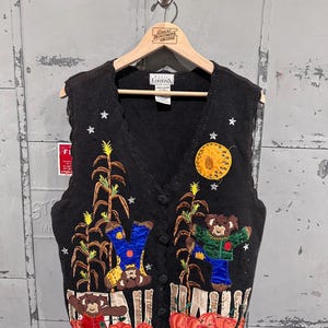 May include: Black knit vest with a V-neck and button closure. The vest features embroidered autumn-themed designs, including scarecrows, pumpkins, corn stalks, a full moon, and stars. The vest is hanging on a wooden hanger.