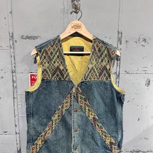 Może przedstawiać: Jeansowa kamizelka z geometrycznym wzorem na karczku i wykończeniach. Kamizelka ma żółtą podszewkę i zapinana jest na guziki z przodu. Wzór zawiera odcienie zieleni, brązu i beżu. Kamizelka wisi na drewnianym wieszaku.