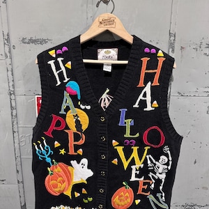 May include: Black knit Halloween vest with colourful embroidered letters spelling "HAPPY HALLOWEEN". The vest features pumpkins, ghosts, and a skeleton design. The vest is button-down and has a decorative orange and white trim at the bottom.