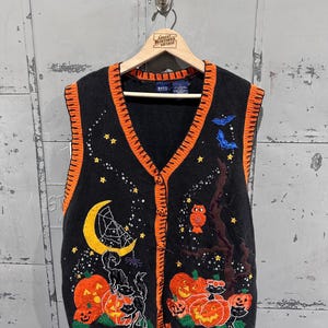 Puede incluir: Chaleco negro con temática de Halloween, con ribete naranja y cierre de botones. Presenta bordados de calabazas, gatos, murciélagos, un búho, una luna creciente y estrellas. El chaleco está colgado de una percha de madera.