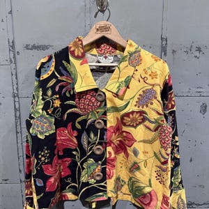 Può includere: Una camicia vintage con bottoni e design diviso. Un lato è nero con motivi floreali rossi e verdi, l'altro è giallo con motivi floreali simili. La camicia ha grandi bottoni di legno ed è appesa a una gruccia di legno.