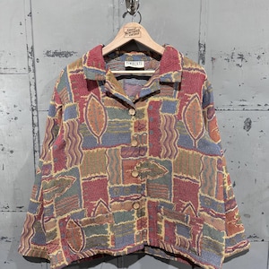 Könnte beinhalten: Vintage-Knopfjacke mit farbenfrohem Patchwork-Design. Die Jacke hat eine Mischung aus roten, blauen, grünen und gelben geometrischen Formen und Blättermustern. Auf dem Etikett steht "TIMBUKTU". Die Jacke hängt an einem Holzkleiderbügel.