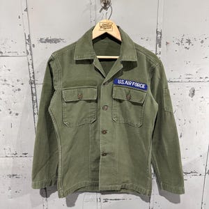 May include: Olive green U.S. Air Force jacket with a blue patch on the chest. The jacket has two chest pockets, button closures, and long sleeves. The jacket is hanging on a wooden hanger.