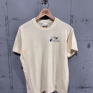 May include: A cream-colored short-sleeve t-shirt featuring a small graphic on the left chest. The graphic depicts a helicopter above the text "FLIGHT TRAILS HELICOPTERS, INC." with a large blue letter F positioned to the left of the text.