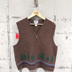 May include: A brown sleeveless knit vest featuring a full-length front zipper and a V-neckline. The lower portion of the vest displays an embroidered pattern of green pine trees above wavy blue and dark navy horizontal lines.