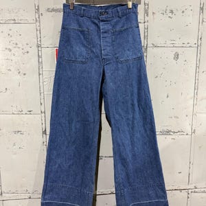 May include: Blue denim pants with a wide leg design. The pants have two front pockets and a button closure. The hemline features a white stitch detail. The pants are hanging on a wooden hanger.