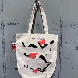 May include: Off-white canvas tote bag with black and red graphic design. The tote features the text "THE NEW YORKER" at the top. The graphic depicts figures reading books. The bag has two long handles.