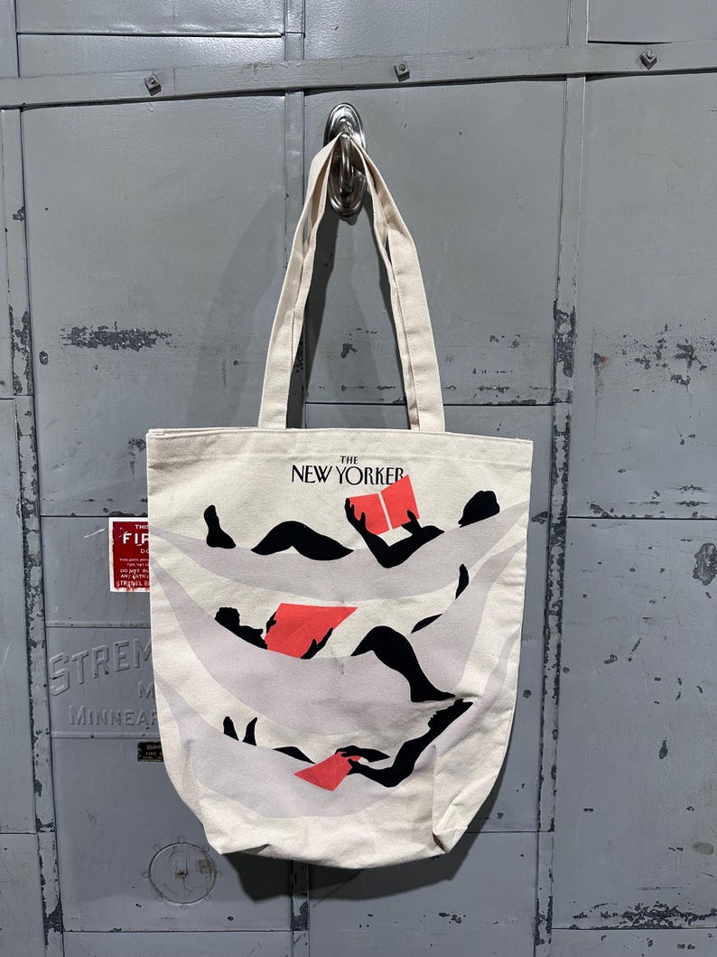May include: Off-white canvas tote bag with black and red graphic design. The tote features the text "THE NEW YORKER" at the top. The graphic depicts figures reading books. The bag has two long handles.