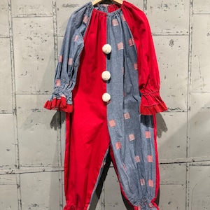 Puede incluir: Un disfraz de payaso vintage, rojo y azul, colgado de una percha de madera. El disfraz está dividido por la mitad, con un lado rojo y el otro azul. Pompones blancos adornan el frente y encaje rojo adorna los puños y tobillos.