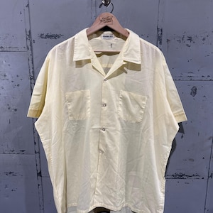 May include: Pale yellow short-sleeved shirt with two front pockets and a button-down closure. The shirt is hanging on a wooden hanger with the text "Great Northern Vintage". The shirt has a relaxed fit and a classic collar.