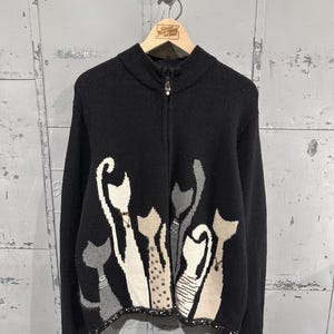 Könnte beinhalten: Ein schwarzer Strickpullover mit Reißverschluss und einem grafischen Muster aus stilisierten Katzen in Weiß-, Grau- und Beigetönen. Der Pullover hat einen Stehkragen, lange Ärmel und einen Saum, der mit kleinen Metallnieten verziert ist.