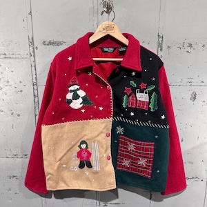 以下が含まれることがあります： 赤、黒、ベージュ、緑色のヴィンテージウィンタージャケット。お祝いをテーマにした刺繍が施されています。雪だるま、スキーヤー、家、雪の結晶が特徴です。ボタン留めで、木製のハンガーに掛けられています。