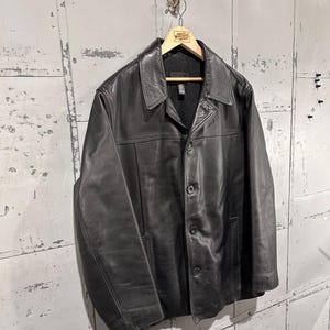 May include: A black leather jacket featuring a classic collar, a button-front closure with three visible buttons, long sleeves, and two side pockets. The jacket is lined with a dark material and is displayed on a wooden hanger.