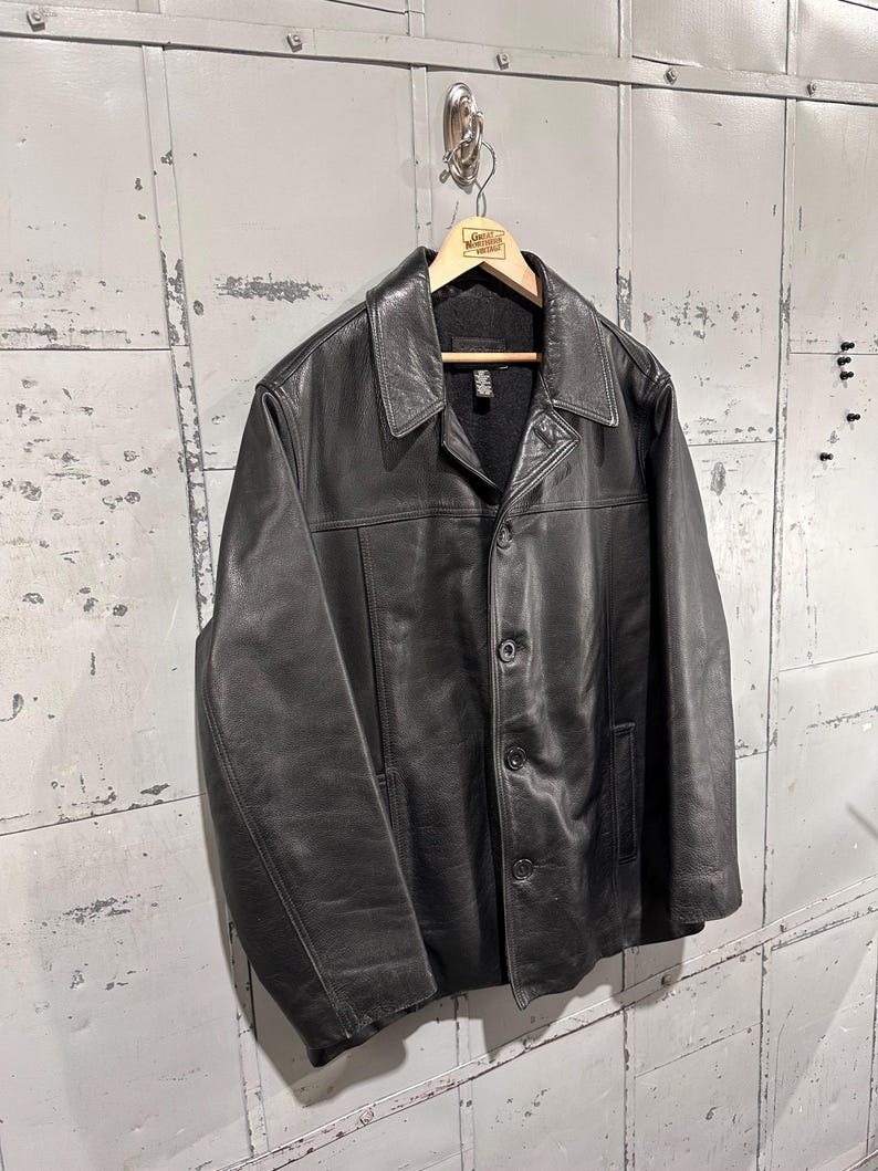 May include: A black leather jacket featuring a classic collar, a button-front closure with three visible buttons, long sleeves, and two side pockets. The jacket is lined with a dark material and is displayed on a wooden hanger.