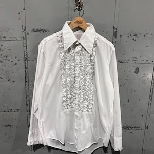 May include: White long-sleeve shirt with a ruffled front. The shirt has a pointed collar and is hanging on a wooden hanger. The shirt is made of a lightweight fabric and has buttoned cuffs.
