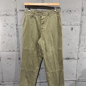 May include: Olive green pants with a relaxed fit, hanging on a wooden hanger. The pants have a button and zipper closure, with multiple pockets and reinforced stitching details. The fabric appears to be a durable cotton blend.
