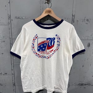 May include: A white ringer t-shirt with dark blue trim on the collar and sleeves. The front features a graphic with the text "FARMFEST 76" and "SEPTEMBER 13-19, 1976 LAKE CRYSTAL MINNESOTA" surrounded by a circular border of red and blue stars.