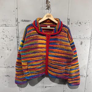 May include: A colorful, vintage, knitted jacket with a red collar and zipper. The jacket features horizontal stripes in various colors, including red, orange, yellow, blue, and green. It is hanging on a wooden hanger against a gray metal background.