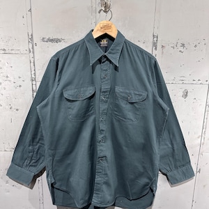 Può includere: Camicia a maniche lunghe color teal con due tasche sul petto. La camicia è appesa a una gruccia di legno con la scritta "Great Northern Vintage". Ha un colletto classico e polsini abbottonati.