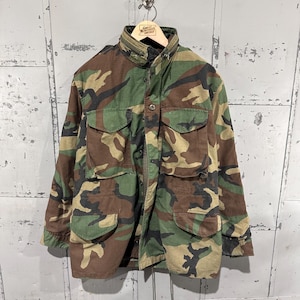 May include: A field jacket featuring a woodland camouflage pattern in shades of green, brown, and black. The jacket includes a high collar with a zipper, a front button closure, two chest pockets with flaps, and two lower pockets with flaps.