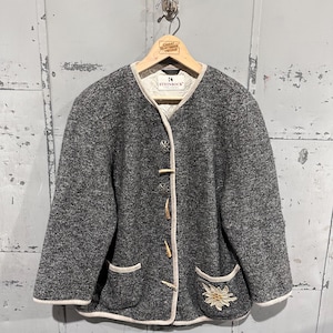 May include: A gray wool jacket with a cream-colored trim and wooden toggle closures. The jacket features two front pockets and a floral embroidery detail on the lower right side. The label reads "STEINBOCK".