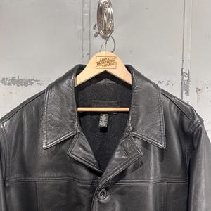 May include: A black leather jacket featuring a wide collar, a button-front closure, and a dark gray interior lining. The jacket includes a visible interior tag with care instructions and is displayed on a wooden hanger with the text Great Northern Vintage.