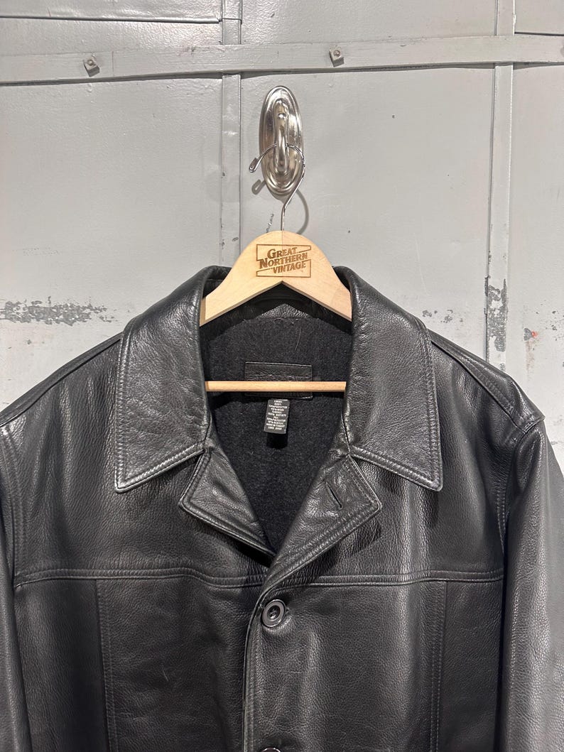 May include: A black leather jacket featuring a wide collar, a button-front closure, and a dark gray interior lining. The jacket includes a visible interior tag with care instructions and is displayed on a wooden hanger with the text Great Northern Vintage.