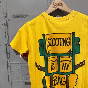 May include: Yellow t-shirt with a graphic of a green and brown backpack. The backpack design includes the words "SCOUTING IS MY BAG" in black lettering. The shirt is hanging on a wooden hanger.