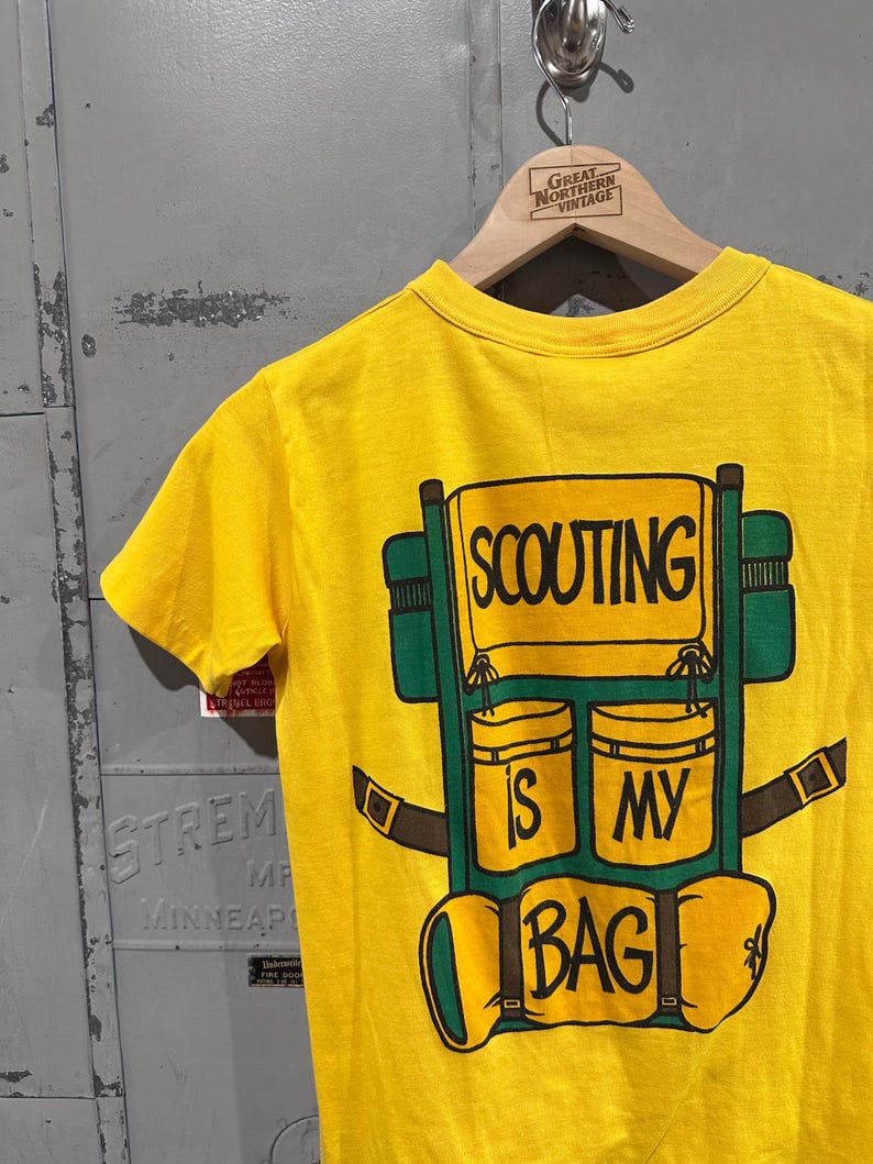 May include: Yellow t-shirt with a graphic of a green and brown backpack. The backpack design includes the words "SCOUTING IS MY BAG" in black lettering. The shirt is hanging on a wooden hanger.