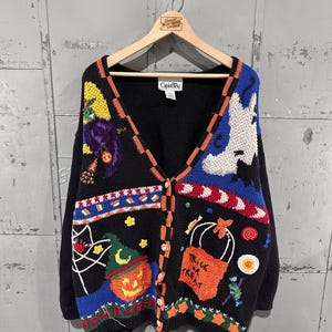 Könnte beinhalten: Eine schwarze Strickjacke mit V-Ausschnitt und bunten Halloween-Motiven wie einer Hexe, einem Geist, einem Kürbis und einer Süßigkeitentüte. Die Knopfleiste ist orange-schwarz kariert und mit weiteren Applikationen wie Sternen verziert.