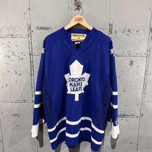 May include: A blue Toronto Maple Leafs hockey jersey featuring a large white maple leaf logo on the chest with the text TORONTO MAPLE LEAFS. The jersey has white horizontal stripes on the sleeves and hem, a V-neck collar, and mesh side panels.