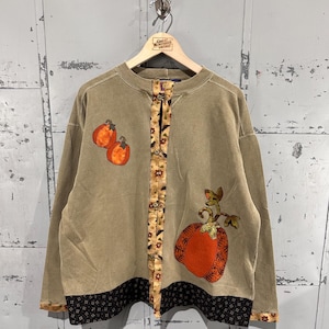 May include: A tan long-sleeve sweatshirt featuring a button-down front placket with a floral pattern, two small orange pumpkin appliqués on the upper right, and a large orange pumpkin appliqué on the lower left with a black patterned hem.
