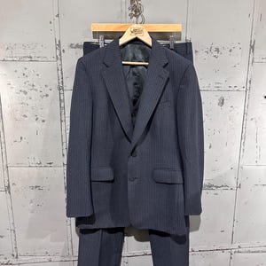 May include: A dark charcoal gray pinstriped suit consisting of a single-breasted blazer with two buttons, a matching vest, and trousers with cuffed hems. The suit is displayed on a wooden hanger, featuring a notched lapel and two flap pockets.