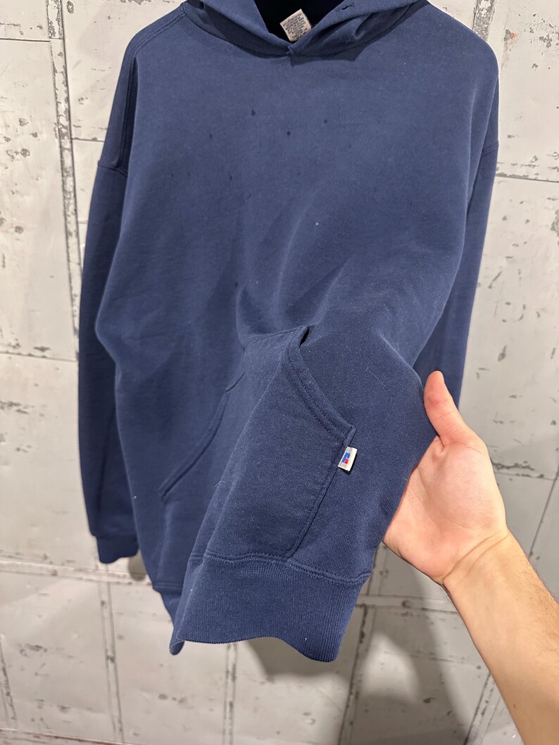 May include: Navy blue hoodie with a kangaroo pocket and a drawstring hood. The sweatshirt has a small white and red tag on the sleeve. The fabric appears to be a soft, medium-weight material.