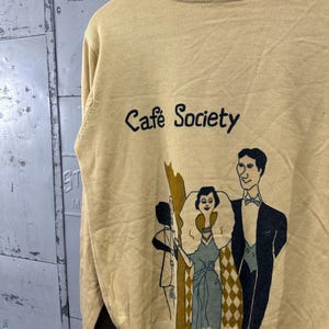 May include: Mustard yellow sweater with the words "Cafe Society" in black script. The sweater features a graphic of three figures in vintage attire. The sweater is hanging on a wooden hanger.