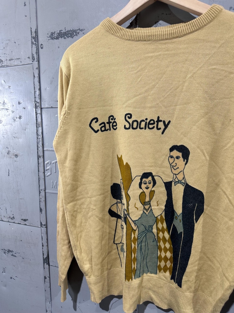 May include: Mustard yellow sweater with the words "Cafe Society" in black script. The sweater features a graphic of three figures in vintage attire. The sweater is hanging on a wooden hanger.