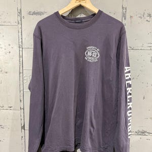 May include: A long-sleeved, purple-grey shirt with the text "PROPERTY OF 86-33 ATHLETICS" on the upper left chest. The right sleeve has the word "ABERCROMBIE" printed vertically in white. The shirt is hanging on a wooden hanger.