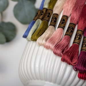 Könnte beinhalten: Eine Sammlung farbenfroher Stickgarne in Blau-, Grün-, Rosa- und Rottönen. Die Garne sind auf einer weißen, gerippten Keramikvase angeordnet, mit Eukalyptusblättern im Hintergrund. Jede Spule ist mit dem Markennamen "DMC" versehen.