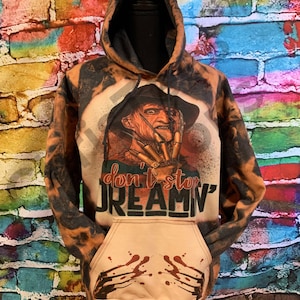 K&ouml;nnte beinhalten: Ein mit Bleichmittel behandelter Kapuzenpullover mit einer Grafik einer Figur mit Klauenhand und Hut. Der Text lautet "don't stop DREAMN'". Der vordere Taschenbereich zeigt eine Grafik von Kratzspuren mit roten Spritzern und eine Kapuze.