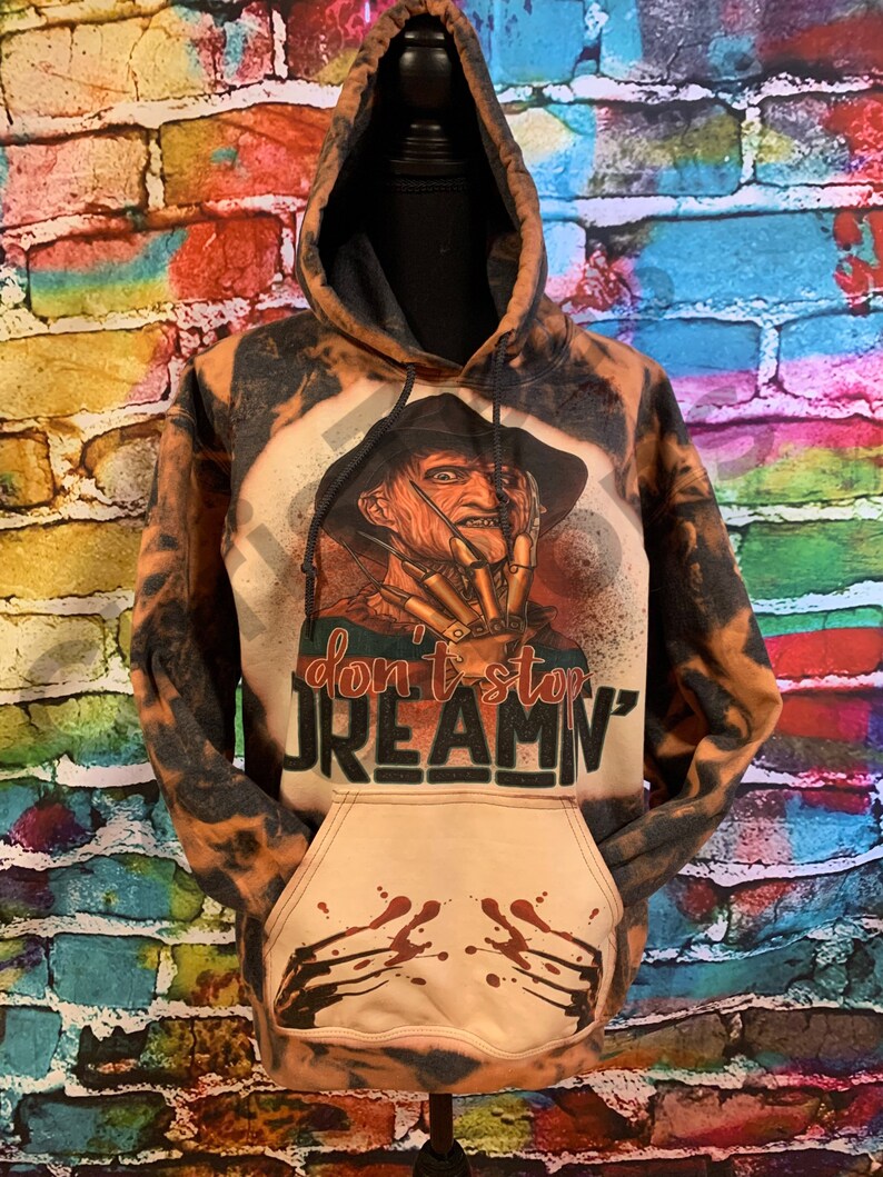 K&ouml;nnte beinhalten: Ein mit Bleichmittel behandelter Kapuzenpullover mit einer Grafik einer Figur mit Klauenhand und Hut. Der Text lautet "don't stop DREAMN'". Der vordere Taschenbereich zeigt eine Grafik von Kratzspuren mit roten Spritzern und eine Kapuze.
