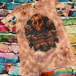May include: A tie-dye t-shirt featuring a graphic of a hockey mask with the text "WELCOME TO CAMP CRYSTAL LAKE". The shirt has a distressed look with blood splatter and a hockey mask on the sleeves.