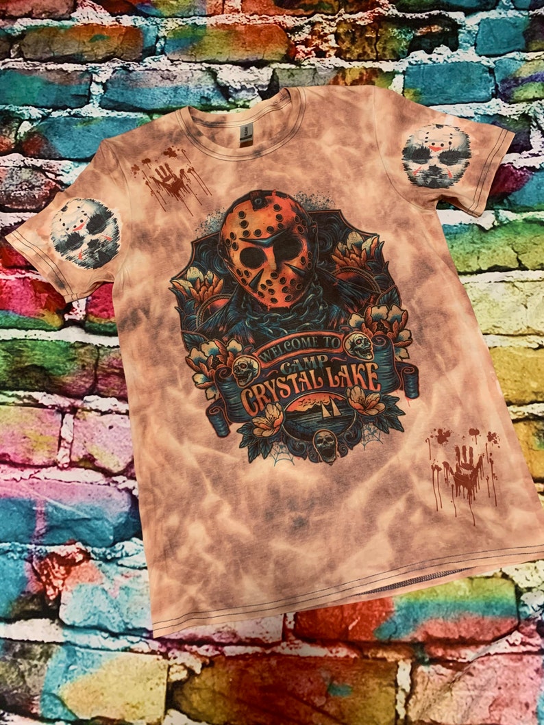 May include: A tie-dye t-shirt featuring a graphic of a hockey mask with the text "WELCOME TO CAMP CRYSTAL LAKE". The shirt has a distressed look with blood splatter and a hockey mask on the sleeves.